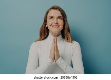 Headshot portrait of happy young woman looking up making wish while folded hands in prayer gesture asking god for help or forgiveness, looking up with smile, isolated on blue studio background