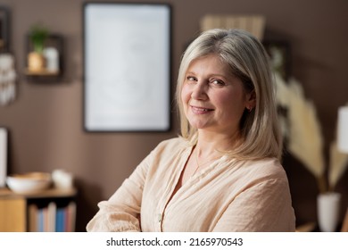 Headshot portrait positive mature woman with healthy smile standing on background of cozy living room, happy middle-aged lady looking to camera, posing for photo at home, enjoying leisure time.
