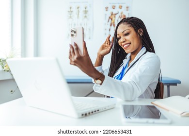 Headshot Portrait Of Positive Smiling African Amerian Woman Doctor In Uniform Sitting At Desk Videocalling At Camera Saying Hi Waving Hand Gesturing Hello. Webcam View. 