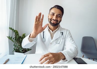 Headshot Portrait Of Positive Smiling Indian Man Doctor In Uniform Sitting At Desk Videocalling At Camera Saying Hi Waving Hand Gesturing Hello. Webcam View. Telemedicine, Online Medicine And E-health