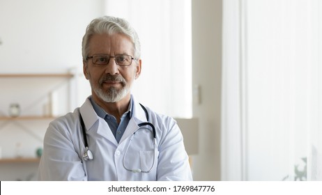 Headshot Portrait Of Serious Mature Male Doctor Or Therapist In White Medical Uniform, Glasses And Stethoscope, Mature Old Man GP Or Physician In Spectacles Pose At Workplace, Healthcare Concept