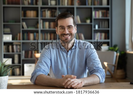 Headshot portrait of smiling 30s Caucasian millennial man in glasses sit at desk at home talk speak on video call or webcam virtual conference. Picture of happy young male in spectacles pose at table.