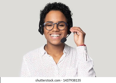 Headshot portrait of smiling african American female call center agent in glasses wearing headset with microphone, happy biracial girl dispatcher in earphones laugh isolated on grey studio background