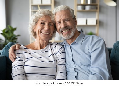 Headshot Portrait Of Smiling Elderly 60s Husband And Wife Sit Relax On Couch Hugging Cuddling, Happy Mature Old Couple Rest On Sofa In Living Room Embrace Look At Camera Show Love And Care