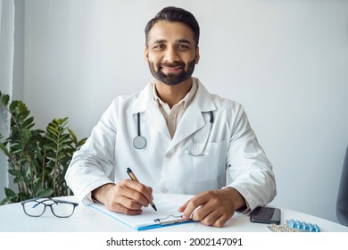 Headshot portrait of smiling indian man doctor working with prescription, choosing right treatment method and medical diagnosis sitting at desk with patient card and pills drugs. Medicine and health - Powered by Shutterstock