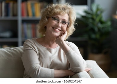 Headshot portrait of smiling middle-aged Caucasian woman in glasses sit on couch in living room relaxing, happy senior female in spectacles rest on sofa at home, look at camera posing, enjoy weekend