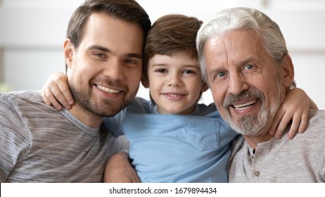 Headshot Portrait Of Smiling Three Generations Of Men Hug Posing For Picture Together At Home, Happy Little Boy Embrace Young Father And Elderly Grandfather Look At Camera Relax In Living Room