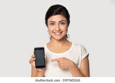 Headshot portrait of smiling young Indian woman isolated on grey studio background point at cellphone screen good online deal. Picture of happy multiracial female user show promotion on smartphone.