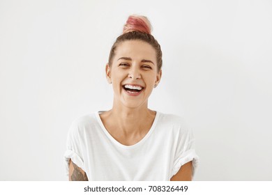 Headshot Of Positive Emotional Gorgeous Young Woman Wearing Pinkish Hair Bun Grinning Broadly, Enjoying Good Day. Beautiful Girl With Tattoo On Arm Having Fun Indoors, Laughing At Something Funny