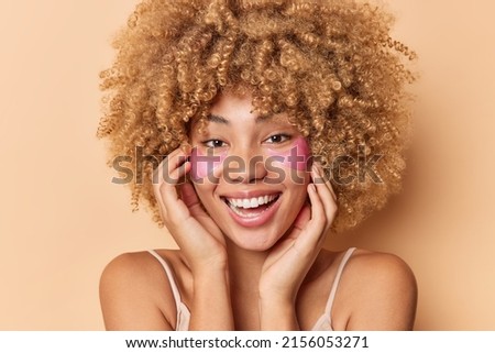 Headshot of positive young curly woman touches face smiles broadly enjoys softness of skin applies pink hydrogel patches reduces puffiness stands bare shoulders isolated over beige background