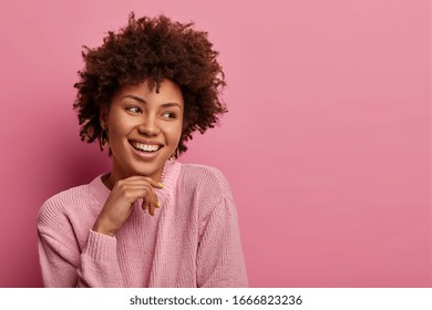 Headshot Of Pretty Charming Curly Afro Woman Poses Joyful And Looks Aside, Keeps Hand Under Chin, Wears Warm Sweater, Stands With Broad Smile, Isolated On Pink Background, Blank Space Aside.