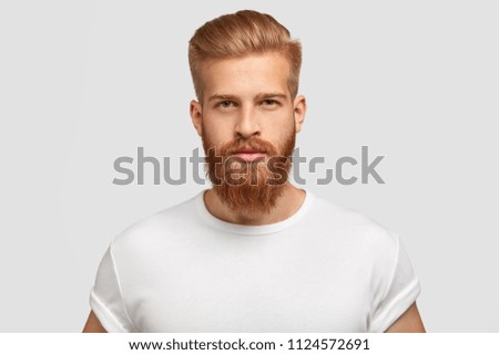 Headshot of red haired serious young male blogger looks confidently at camera, thinks about new content of his web page, dressed casually, earns money distantly, isolated over white background