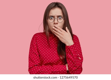 Headshot Of Scared Young Woman Covers Mouth With Hand, Has Frightened Puzzled Facial Expression, Dressed In Polka Dot Blouse, Sees Something Horrible, Isolated Over Pink Background. Omg Concept