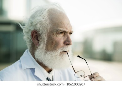 Headshot Of Senior Doctor Wearing White Coat Looking Away While Wrapped Up In Thoughts, Blurred Background