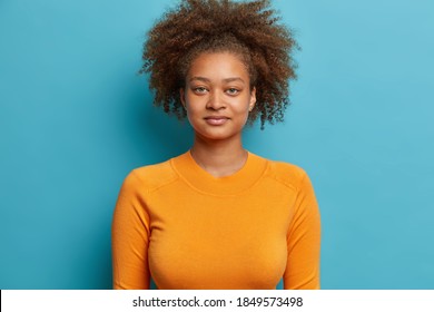 Headshot of serious curly haired teenager girl looks satisfied at camera dreessed in casual orange jumper poses against blue background. Pretty young Afro American female with bushy wavy hair