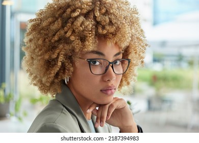 Headshot of serious successful woman with curly blonde hair keeps hand under chin wears optical spectacles and formal clothes looks attentively at camera poses outdoors against blurred background