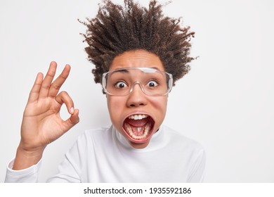 Headshot of shocked emotive curly haired young woman stares at camera has jaw dropped raised hair shows okay gesture says I like it has crazy look isolated over white background. Madness concept