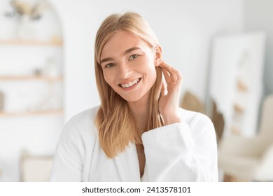 Headshot of smiling blonde woman in white robe posing gently touching her hair and face, enjoying skincare beauty routine at home bathroom, looking at camera. Self pampering concept