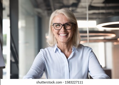 Headshot of smiling middle-aged businesswoman in glasses look at camera pose shooting live webinar broadcast in office, happy mature female employee talk on video call at workplace, leadership concept