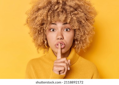 Headshot of surprised curly haired young woman presses index finger over lips makes shush gesture looks mysteriously at camera wears jumper isolated over vivid yellow background tells secret
