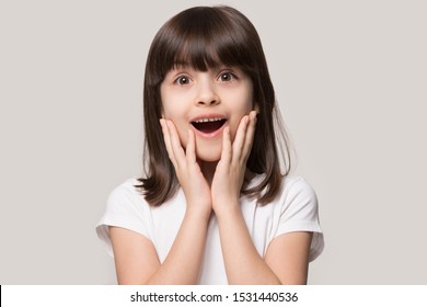 Headshot of surprised little preschooler girl look at camera feel stunned amazed by unexpected good news, happy small child isolated on grey studio background shocked by unbelievable surprise