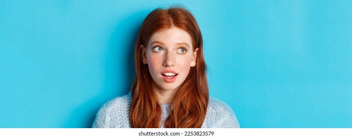 Headshot of thoughtful redhead teen girl looking at upper left corner, staring at logo with curious expression, standing over blue background. - Shutterstock ID 2253823579