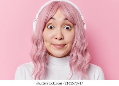 Headshot of young Asian woman with pink hair looks with widely opened eyes feels amazed wears white turtleneck finds out shocking news listens audio track via headphones isolated over rosy wall