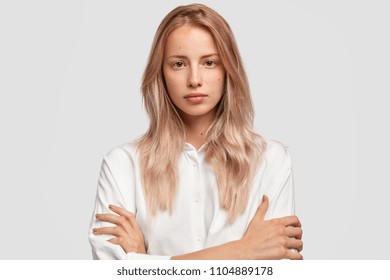 Headshot Of Young Caucasian Female With Serious Expression, Has Healthy Pure Skin, Keeps Arms Folded, Has Long Straight Hair, Listens Attentively Necessary Information, Stands Against White Wall