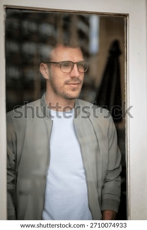 Headshot of a young man standing at the window and looking thoughfully. Confident man wearing white shirt and jacket. Photographed through the window. 