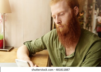 Headshot Of Young Redhead Male Wearing Green Casual Shirt, Holding Generic Cell Phone, Browsing Internet And Chatting With Friends On Electronic Device, Sitting Against Cafe Interior Background