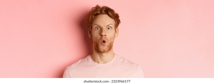 Headshot of young redhead man with beard, saying wow and staring at camera amazed, checking out promo deal, standing over pink background.