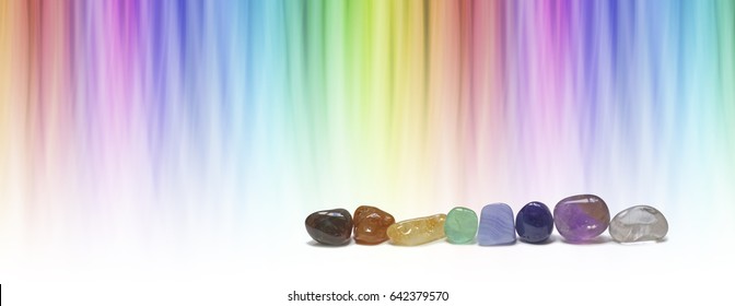 Healing chakra crystals and color healing website header -  A  row of eight chakra colored tumbled crystals and against a linear graduated chakra rainbow color background with  copy space

