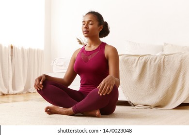 Health, active lifestyle, well being and people concept. Portrait of attractive peaceful young black dark-skinned female in stylish sports clothes meditating indoors, keeping her eyes closed