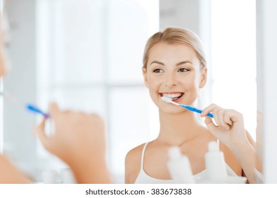 Health Care, Dental Hygiene, People And Beauty Concept - Smiling Young Woman With Toothbrush Cleaning Teeth And Looking To Mirror At Home Bathroom