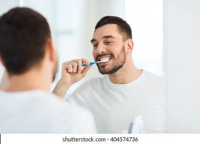 health care, dental hygiene, people and beauty concept - smiling young man with toothbrush cleaning teeth and looking to mirror at home bathroom