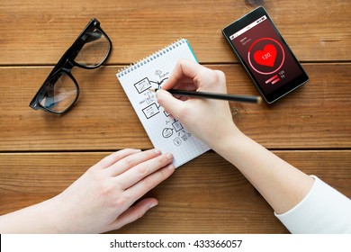 Health Care, Planning And People Concept - Close Up Of Woman Or Student Drawing Scheme In Notepad With Pencil And Eyeglasses With Heart Rate On Smartphone On Wooden Table