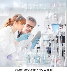 Health Care Researchers Working In Life Scientific Laboratory. Young Female Research Scientist And Senior Male Supervisor Looking Focused At Tablet Computer Screen Evaluating And Analyzing Study Data.