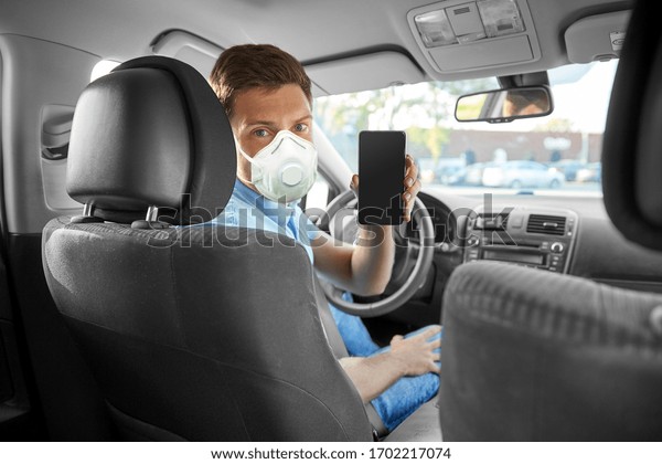 health care, safety and pandemic\
concept - man or car driver wearing face protective mask or\
respirator for protection from virus disease showing\
smartphone