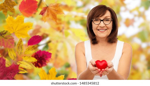 Health, Charity And Valentine's Day Concept - Portrait Of Smiling Senior Woman Holding Red Heart Over Autumn Leaves And Nature Background