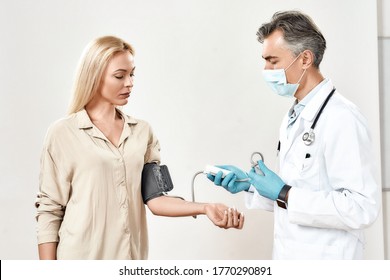 Health Check Up. Male Mature Doctor Wearing Protective Face Mask And Blue Sterile Gloves Measuring Blood Pressure Of Young Woman, His Female Patient. Healthcare And Medicine. Medical Consultation