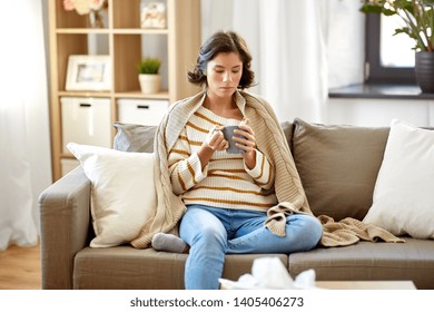 Health, Cold And People Concept - Sad Sick Young Woman In Blanket Sitting On Sofa And Drinking Hot Tea At Home