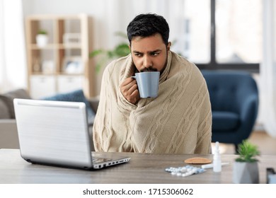 Health, Cold And People Concept - Sick Young Indian Man In Blanket With Laptop Computer Drinking Hot Tea And Working At Home