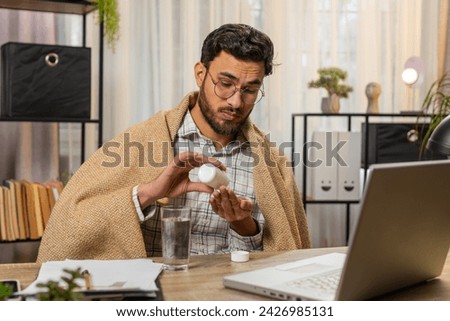 Health, cold, and people concepts. Sick young Hispanic businessman wrapped in blanket works on laptop at home office desk. Unhealthy Hispanic freelancer takes medicine, shivering from the cold at home