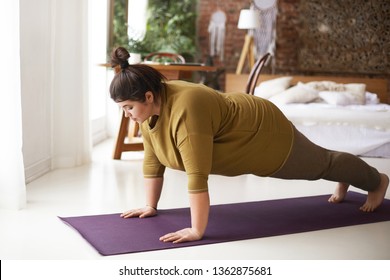 Health, Fitness And Sports Concept. Plus Size Young Brunette Woman Doing Plank Exercise On Mat In Cozy Bedroom Interior, Going To Loose Extra Pounds, Become Strong And Fit, Training Endurance