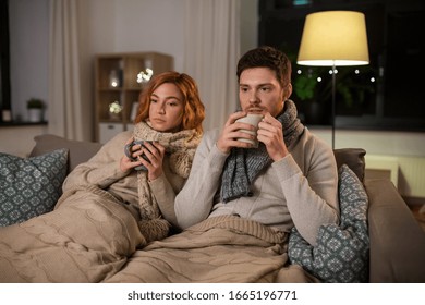 Health, Flu And People Concept - Sick Young Couple In Scarves Drinking Hot Tea At Home