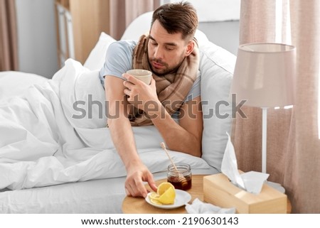 health, illness and people concept - sick man drinking hot tea with lemon and honey in bed at home