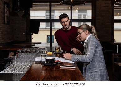 Health inspector sitting in a restaurant and writes a fine and a negative report to the owner or manager of the cafeteria after she finds poor hygiene conditions in the kitchen. Sanitary inspection - Shutterstock ID 2277721479