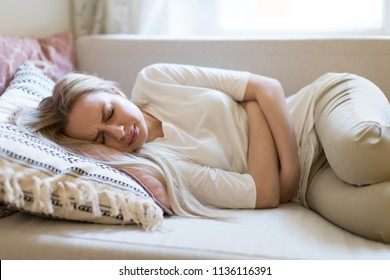 Health issues problems.Young Caucasian woman suffering from stomach pain, feeling abdominal pain or cramps, lying on sofa.Period menstruation, female health problem, aching belly and gynecology - Shutterstock ID 1136116391
