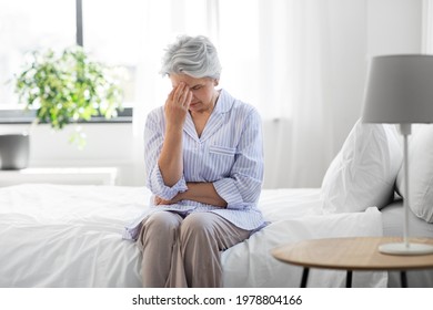 Health, Old Age And People Concept - Senior Woman In Pajamas Suffering From Headache Sitting On Bed At Home Bedroom