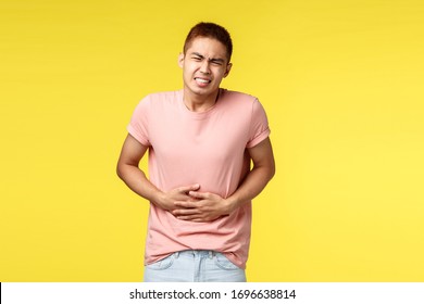 Health, people and food concept. Asian guy in pink t-shirt, feel sick, grimacing from pain in stomach, standing over yellow background suffer from poisoning, hold hands on belly and bending